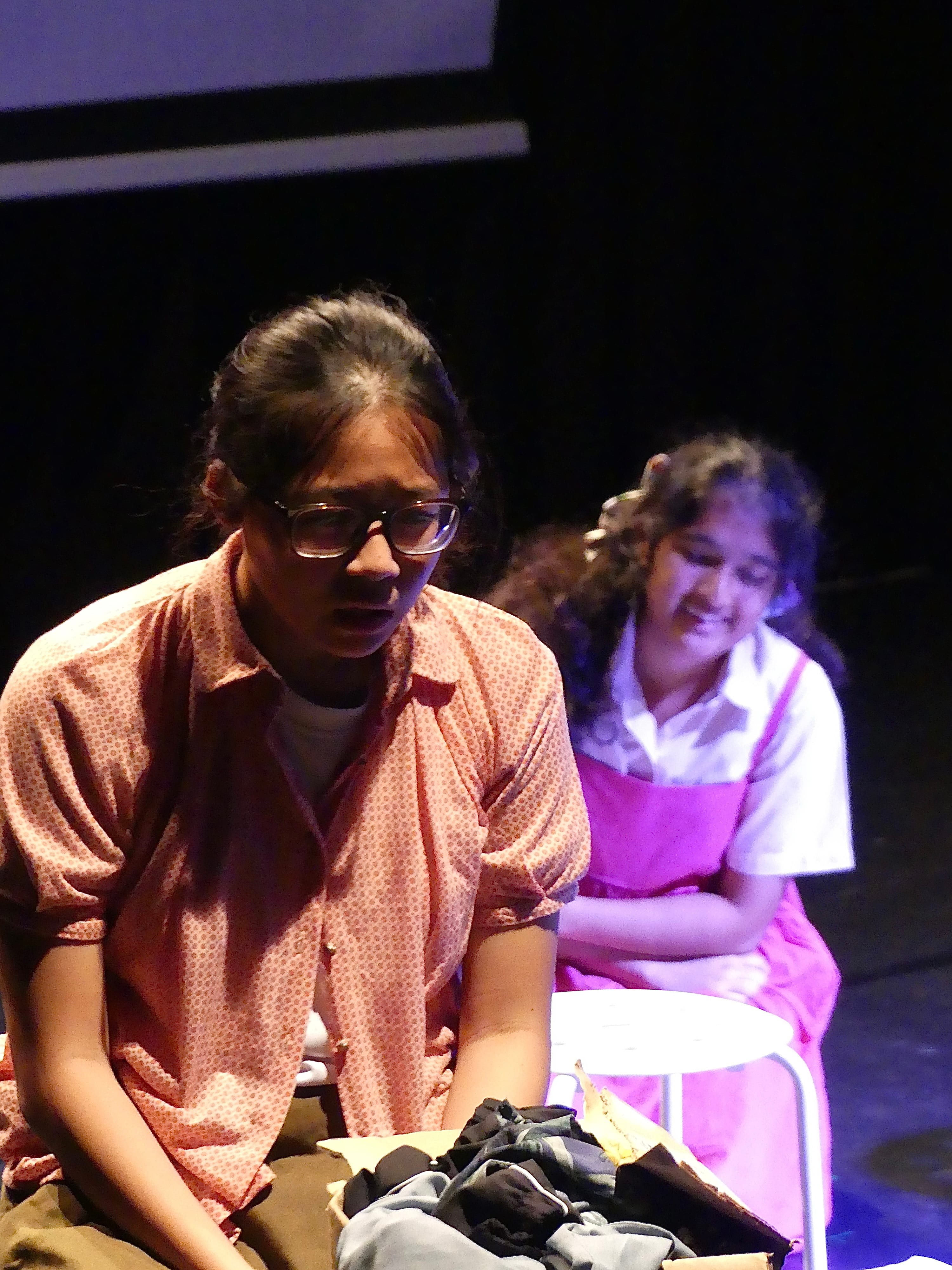 Two young female students perform on a darkened stage, one in a pink floral shirt delivering lines expressively in the foreground, the other in a pink dress smiling in the background.